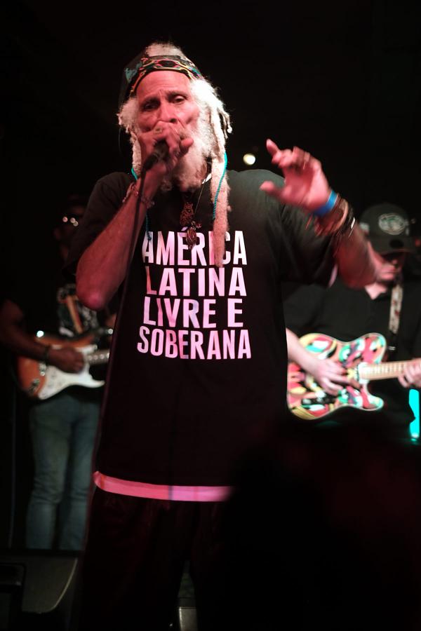 Cedric Myton of The Congos performing on stage wearing a black t-shirt with pink text reading “AMERICA LATINA LIVRE E SOBERANA” (Free and Sovereign Latin America). He holds a microphone and wears a colorful headband with white dreadlocks visible, his arm raised energetically.