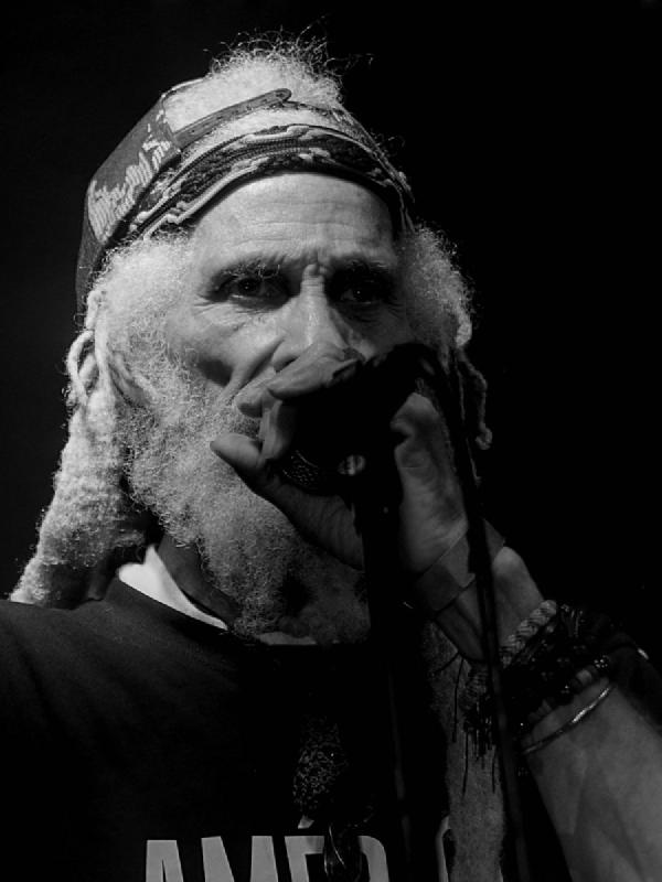 Black and white close-up portrait of Cedric Myton performing, wearing a cap with white curly hair and beard visible. He holds a microphone close to his mouth with an intense, passionate expression while performing.
