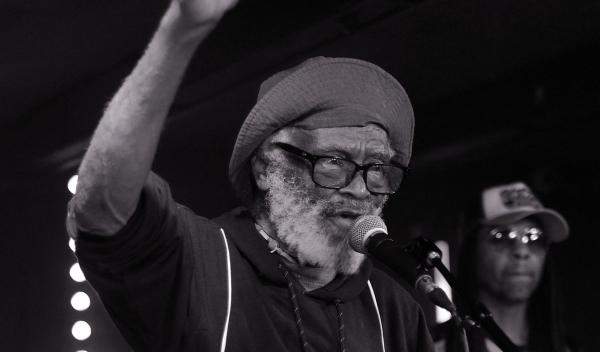 Black and white photograph of Watty Burnett performing live on stage. The veteran reggae artist wears glasses and a beanie hat while singing into a microphone with one arm raised expressively. Stage lights are visible in the background.