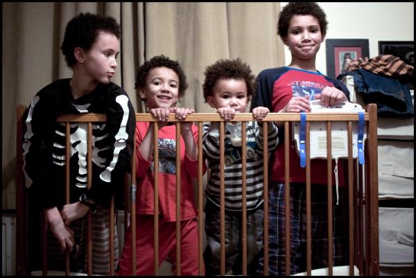 Four young children of varying ages standing behind a wooden baby cot or crib with vertical slats. The children are wearing casual clothing including striped and colorful tops, and they appear to be in a home setting with cream-colored walls.