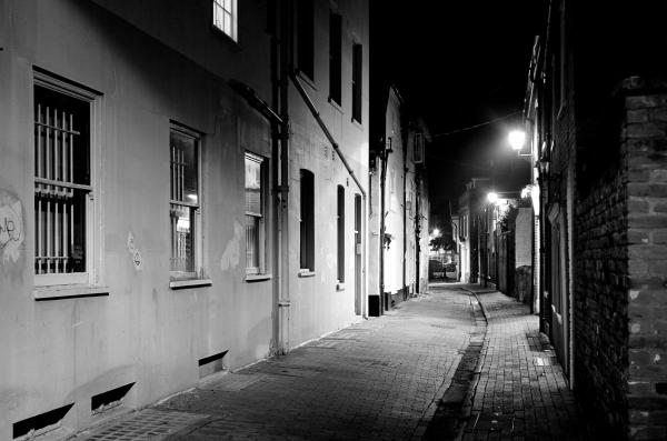 Black and white night photograph of a narrow cobblestone alley with weathered buildings on both sides, illuminated by street lamps creating pools of light in the darkness
