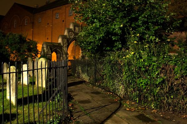 Night photograph of a historic brick church with illuminated archway entrance, viewed through a cemetery gate with gravestones visible in the foreground and overgrown vegetation framing the scene