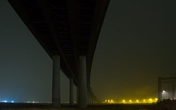 Atmospheric night photograph beneath a large bridge or viaduct, showing massive concrete support pillars silhouetted against a misty sky with distant lights glowing in amber tones