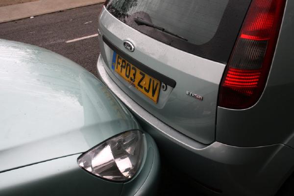 A silver car is parked with its rear bumper pressed up against the front bumper of another light green car on the street, showing no visible damage. Both vehicles are wet from rain, with the silver car’s number plate “FP03 ZJV” clearly visible.
