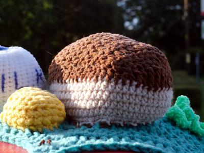 A close-up view of crocheted food items sitting on the post box's yarn-bombed surface. The scene includes a white milk bottle with MILK lettered in blue, a brown loaf of bread, a yellow knob of butter and various other crocheted food elements arranged on a turquoise textured base. These detailed food replicas showcase the intricate craftsmanship typical of modern UK postbox and telephone box yarn bombing installations.