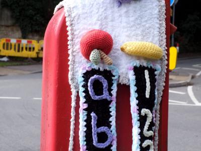 A red British post box covered in elaborate white and purple crocheted yarn bombing art. On top sits a crocheted owl wearing a graduation cap with blue eyes, purple scarf, and multicolored clothing. The phone box displays the letters ABC and 123 in purple lettering on white crocheted panels, with decorative mushrooms red toadstool and yellow varieties adorning the sides. The entire structure is wrapped in white textured yarn work with purple trim, representing a whimsical example of UK yarn bombing street art.