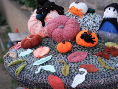 A detailed view of a knitted Halloween tableau showing multiple crocheted pumpkins of varying sizes and colors including orange, pink, white, and patterned varieties. A black cat in a witch's hat sits among the pumpkins, with one orange pumpkin featuring a black silhouette design. The display includes autumn leaves in various colors (orange, red, white, yellow, green) scattered across a multi-colored knitted base with geometric patterns. The scene is photographed outdoors with stone architecture visible in the background.