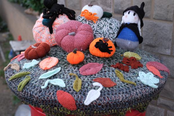 A detailed view of a knitted Halloween tableau showing multiple crocheted pumpkins of varying sizes and colors including orange, pink, white, and patterned varieties. A black cat in a witch&rsquo;s hat sits among the pumpkins, with one orange pumpkin featuring a black silhouette design. The display includes autumn leaves in various colors (orange, red, white, yellow, green) scattered across a multi-colored knitted base with geometric patterns. The scene is photographed outdoors with stone architecture visible in the background.