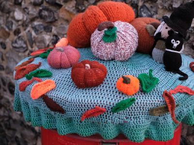 A colorful knitted Halloween display featuring various crocheted pumpkins in orange, red, and pink arranged on a turquoise knitted tablecloth. The display includes a black cat figure wearing a witch's hat, autumn leaves in red and orange, and small decorative elements scattered around. The tablecloth has a scalloped edge and the scene is photographed outdoors against a stone wall background.