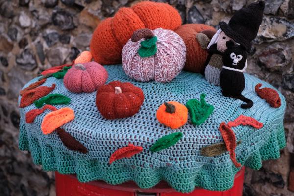 A colorful knitted Halloween display featuring various crocheted pumpkins in orange, red, and pink arranged on a turquoise knitted tablecloth. The display includes a black cat figure wearing a witch&rsquo;s hat, autumn leaves in red and orange, and small decorative elements scattered around. The tablecloth has a scalloped edge and the scene is photographed outdoors against a stone wall background.
