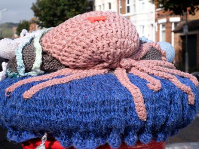 A knitted blue and pink octopus sits atop a red postbox, with multiple tentacles draping over the sides and buildings along a street visible behind.