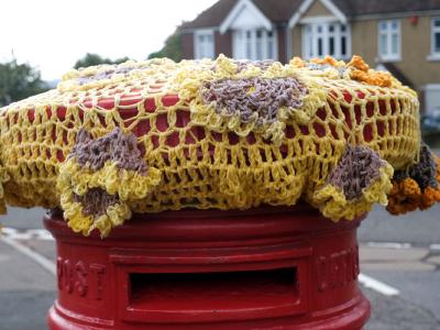 A red UK postbox is topped with a handmade crochet cover in yellows and browns, featuring sunflower and petal designs, with suburban houses in the background.