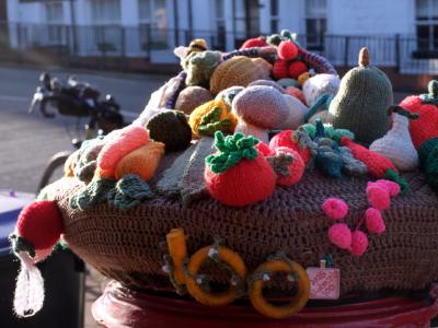 A large knitted and crocheted harvest sculpture featuring colorful wool vegetables and fruits displayed outdoors in a residential street
