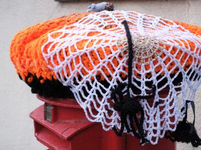 Crocheted Halloween decoration in the form of an orange pumpkin with white mesh netting draped over a traditional red British Royal Mail postbox, complete with a grey knitted stem and black decorative elements