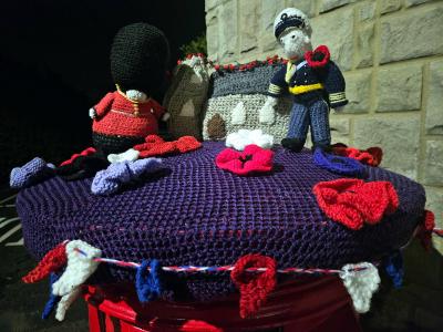 A handcrafted Remembrance Day tribute featuring knitted figures and elements arranged on a purple knitted postbox topper. The scene includes a British Queen's Guard soldier in red uniform with black bearskin hat on the left, a miniature knitted cenotaph monument in the center draped with a red poppy garland, and a naval or air force officer figure in navy blue uniform on the right. Multiple red knitted poppies are scattered across the purple surface, along with small red, white, and blue decorative elements. The display sits on a red postbox against a stone wall background.