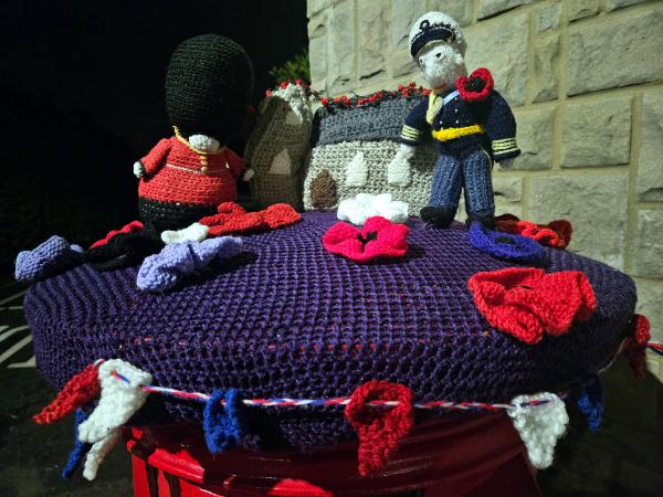 A handcrafted Remembrance Day tribute featuring knitted figures and elements arranged on a purple knitted postbox topper. The scene includes a British Queen’s Guard soldier in red uniform with black bearskin hat on the left, a miniature knitted cenotaph monument in the center draped with a red poppy garland, and a naval or air force officer figure in navy blue uniform on the right. Multiple red knitted poppies are scattered across the purple surface, along with small red, white, and blue decorative elements. The display sits on a red postbox against a stone wall background.