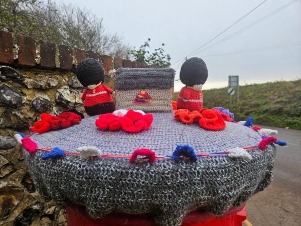 Hand-knitted commemorative display featuring miniature figures of Royal Guards in red uniforms and black bearskin hats positioned behind a knitted memorial stone. The stone is adorned with bright red poppies and sits on a crocheted base decorated with red, white, and blue flowers. The scene is photographed at dawn or dusk against a stone wall backdrop with countryside visible in the distance.