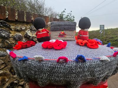 Hand-knitted commemorative display featuring miniature figures of Royal Guards in red uniforms and black bearskin hats positioned behind a knitted memorial stone. The stone is adorned with bright red poppies and sits on a crocheted base decorated with red, white, and blue flowers. The scene is photographed at dawn or dusk against a stone wall backdrop with countryside visible in the distance.