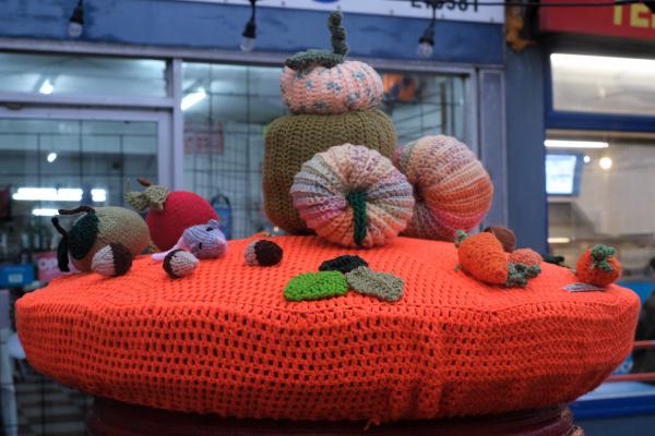 An autumnal post box topper displayed on a large circular bright orange crocheted platform. The centerpiece features a tower of textured knitted pumpkins in various sizes - pink with blue speckles, olive green, and coral - with decorative stems. Scattered around the base are smaller crocheted elements including miniature pumpkins, brown acorns, colorful mushrooms (red and brown), green oak leaves, and small knitted figures. The display is positioned in front of a blue-painted storefront with large windows, creating a festive seasonal scene