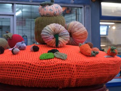 An autumnal post box topper displayed on a large circular bright orange crocheted platform. The centerpiece features a tower of textured knitted pumpkins in various sizes - pink with blue speckles, olive green, and coral - with decorative stems. Scattered around the base are smaller crocheted elements including miniature pumpkins, brown acorns, colorful mushrooms (red and brown), green oak leaves, and small knitted figures. The display is positioned in front of a blue-painted storefront with large windows, creating a festive seasonal scene