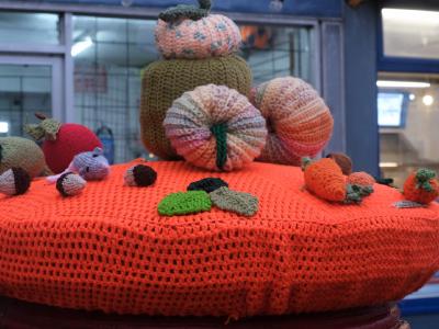 An autumnal post box topper displayed on a large circular bright orange crocheted platform. The centerpiece features a tower of textured knitted pumpkins in various sizes - pink with blue speckles, olive green, and coral - with decorative stems. Scattered around the base are smaller crocheted elements including miniature pumpkins, brown acorns, colorful mushrooms (red and brown), green oak leaves, and small knitted figures. The display is positioned in front of a blue-painted storefront with large windows, creating a festive seasonal scene