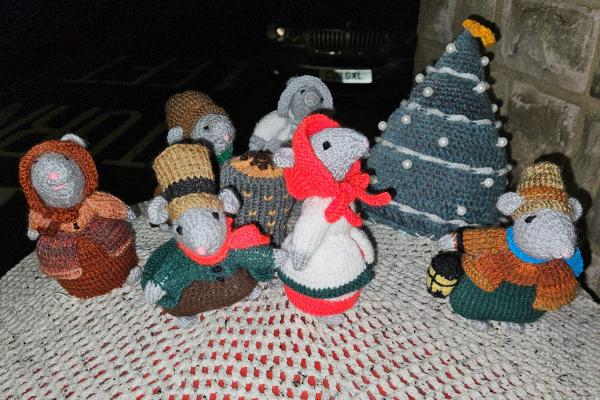 A collection of hand-crocheted festive mice figurines arranged around a decorated Christmas tree on top of a post box in Upper Higham. The display features seven mice dressed in Victorian-style winter clothing including bonnets, coats, and scarves in earthy tones of brown, orange, green, and grey. One central mouse wears a bright red coat and scarf. The dark blue-green crocheted Christmas tree is adorned with white garland spirals, white pompom ornaments, and a yellow star topper. The scene is photographed just before dawn.