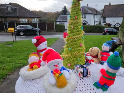 A festive knitted Christmas scene displayed on top of a red Royal Mail post box in a residential street. The handcrafted topper features a winter wonderland tableau on a circular white crocheted base with red detail stitching. At the center stands a tall green spiral Christmas tree decorated with colorful baubles in pink, blue, and other bright colors, topped with a golden yellow star. Surrounding the tree are multiple knitted figures: two white snowmen wearing red Santa hats with white pompoms, one holding a yellow gift, and several small dolls dressed in colorful winter clothing - including figures in red, blue, and green striped outfits with knitted hats. There's also a brown teddy bear character among the group. The scene is photographed outdoors with suburban houses, parked cars, hedges, and a cloudy sky visible in the background, creating a charming community Christmas display.