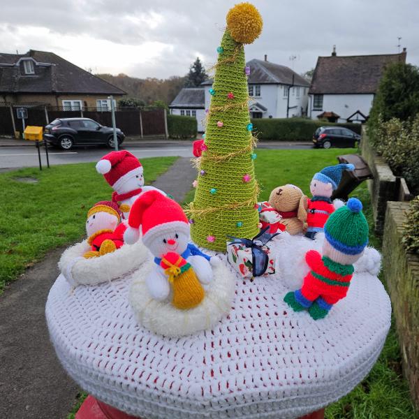 A festive knitted Christmas scene displayed on top of a red Royal Mail post box in a residential street. The handcrafted topper features a winter wonderland tableau on a circular white crocheted base with red detail stitching. At the center stands a tall green spiral Christmas tree decorated with colorful baubles in pink, blue, and other bright colors, topped with a golden yellow star. Surrounding the tree are multiple knitted figures: two white snowmen wearing red Santa hats with white pompoms, one holding a yellow gift, and several small dolls dressed in colorful winter clothing - including figures in red, blue, and green striped outfits with knitted hats. There’s also a brown teddy bear character among the group. The scene is photographed outdoors with suburban houses, parked cars, hedges, and a cloudy sky visible in the background, creating a charming community Christmas display.