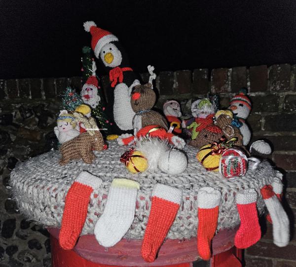 A red pillar postbox at night is covered with a chunky grey knitted topper decorated with handmade festive crochet figures. On top sit a large penguin in a Santa hat and scarf, reindeer, Santas, snowmen, angels and other small Christmas characters, surrounded by knitted baubles and presents, while tiny red-and-white Christmas stocking decorations are stitched all around the edge.
