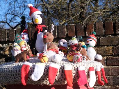 A red pillar postbox at night is covered with a chunky grey knitted topper decorated with handmade festive crochet figures. On top sit a large penguin in a Santa hat and scarf, reindeer, Santas, snowmen, angels and other small Christmas characters, surrounded by knitted baubles and presents, while tiny red-and-white Christmas stocking decorations are stitched all around the edge.