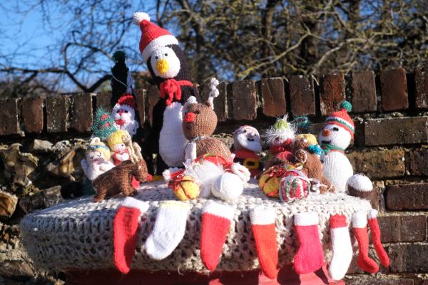 A red pillar postbox at night is covered with a chunky grey knitted topper decorated with handmade festive crochet figures. On top sit a large penguin in a Santa hat and scarf, reindeer, Santas, snowmen, angels and other small Christmas characters, surrounded by knitted baubles and presents, while tiny red-and-white Christmas stocking decorations are stitched all around the edge.