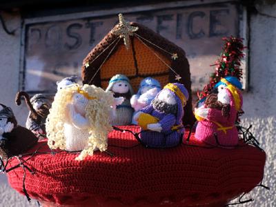 Hand-knitted nativity scene displayed as a festive topper on a village post box, featuring yarn figures of Mary, Joseph, baby Jesus, an angel with curly blond hair, and the three wise men gathered in front of a crocheted stable, with a faded “Post Office” sign on the wall behind.