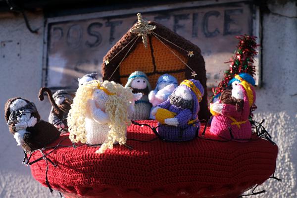 Hand-knitted nativity scene displayed as a festive topper on a village post box, featuring yarn figures of Mary, Joseph, baby Jesus, an angel with curly blond hair, and the three wise men gathered in front of a crocheted stable, with a faded Post Office sign on the wall behind.