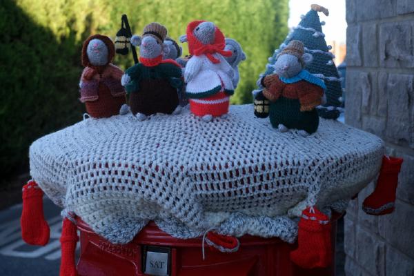A collection of hand-crocheted festive mice figurines arranged around a decorated Christmas tree on top of a post box in Upper Higham. The display features seven mice dressed in Victorian-style winter clothing including bonnets, coats, and scarves in earthy tones of brown, orange, green, and grey. One central mouse wears a bright red coat and scarf. The dark blue-green crocheted Christmas tree is adorned with white garland spirals, white pompom ornaments, and a yellow star topper. The scene is photographed just before dawn.