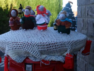A collection of hand-crocheted festive mice figurines arranged around a decorated Christmas tree on top of a post box in Upper Higham. The display features seven mice dressed in Victorian-style winter clothing including bonnets, coats, and scarves in earthy tones of brown, orange, green, and grey. One central mouse wears a bright red coat and scarf. The dark blue-green crocheted Christmas tree is adorned with white garland spirals, white pompom ornaments, and a yellow star topper. The scene is photographed just before dawn.