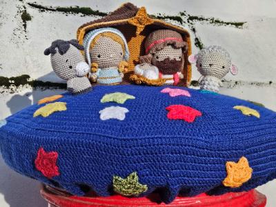 An intricate crocheted nativity scene postbox topper displayed on a red Royal Mail postbox in Ash, Kent, featuring a detailed manger scene with Mary in blue robes holding baby Jesus, Joseph with a red headband, and three amigurumi-style animals (a grey donkey, white sheep, and grey elephant) positioned around the holy family. The figures are set within a golden-brown stable structure against a white weathered wall with moss growth. The entire scene sits atop a rich blue circular crocheted base decorated with colorful sheep motifs in red, orange, yellow, green, white, and pink scattered across the surface, creating a whimsical and heartwarming celebration of the Christmas story through the traditional craft of postbox topper yarn-bombing.