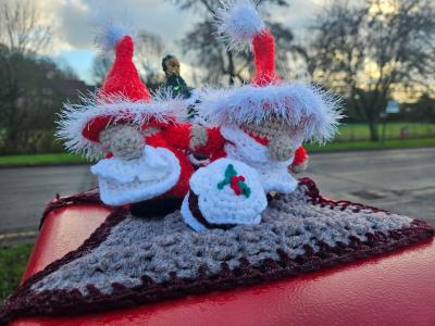A close-up view of a charming crocheted Christmas scene atop a red British postbox, featuring two festive figures in red Santa outfits with white fluffy trim and matching Santa hats topped with sparkly blue tinsel pom-poms. Between them sits a small white crocheted Christmas pudding decorated with red and green holly berries. The figures rest on a grey and burgundy striped knitted base that wraps around the curved top of the postbox. The background shows a residential street with bare winter trees, green grass verges, and a cloudy sky, capturing the whimsical tradition of festive postbox toppers in British communities.