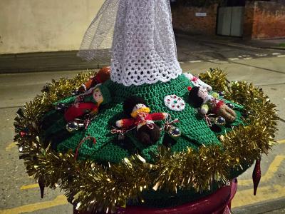 An elegant Christmas postbox topper photographed at night, featuring a tall angel figure with white crocheted wings and a sparkly silver dress standing atop a green knitted base decorated with small festive figures. A doll with blonde hair serves as the angel, dressed in shimmering white and silver garments with outstretched arms. The circular base is adorned with gold tinsel trim around the edge and small crocheted figures including what appear to be carolers or nativity characters, along with decorative elements like a snowman. The scene is set against a dark evening sky with street lights and residential buildings visible in the background.