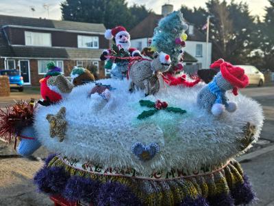 A festive and elaborate crocheted postbox topper featuring a snowy winter wonderland scene with multiple knitted characters creating a bustling Christmas tableau. The white fluffy base, resembling snow, supports various figures including Santa Claus in red, carolers, a grey donkey, birds (including a robin with a red breast), and other festive characters, all arranged around a decorated Christmas tree and holly decoration. The display sits on a multi-layered circular base with white, blue, and textured yarns in various colors, trimmed with tinsel and decorative edging. The scene is photographed during daytime against a residential street backdrop with houses and cars visible.