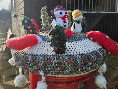A delightful crocheted postbox topper photographed in bright sunlight, featuring an elaborate festive scene on a circular base with red, white, and green striped patterns. The display includes two snowmen figures (one with a colorful striped hat and red scarf, another with a yellow turban-style hat), decorative Christmas trees in various shades of green with tinsel details, a small red stocking, and a dark Christmas tree decoration. In the foreground, a small crocheted Christmas tree with sparkly green yarn and a red star on top takes center stage. Red mittens or stockings accent the edges of the circular base. The postbox topper sits atop a red Royal Mail postbox with white fluffy trim visible at the base, while a black-painted wooden building and garden furniture are visible in the sunny background.
