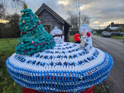 A festive hand-knitted postbox topper displayed on a red British Royal Mail postbox, featuring a charming winter scene with three crocheted snowmen figures and a decorated teal Christmas tree with colorful baubles and a gold star. The scene is set on a circular blue and white crocheted base with intricate striped patterns. The snowmen have white fluffy bodies, orange carrot noses, and happy expressions, with one holding what appears to be a red mitten or scarf. Behind the display, a dark wooden building and residential street are visible under a cloudy winter sky, capturing the community spirit of festive yarn-bombing in a British village.