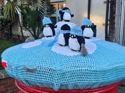 A charming crocheted postbox topper featuring a winter scene with adorable penguin figures on an ice-blue circular base. The display includes five knitted penguins wearing matching light blue hats and scarves, arranged around white crocheted snowballs or igloos on top of a textured pale blue base that resembles ice. The word SOUTH appears in red lettering on the side. A grey crocheted seal sits on the edge. The topper is displayed on a red Royal Mail postbox in front of shop windows showing reflections of buildings, creating a playful Arctic-themed Christmas decoration.