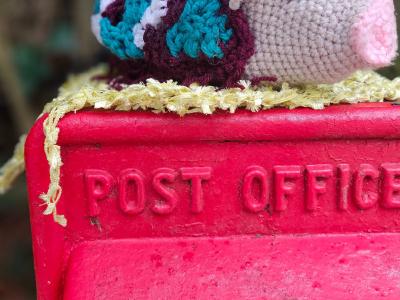 Crocheted multicoloured pig postbox topper with long eyelashes sitting on a red “Post Office” box in Barham, Kent.