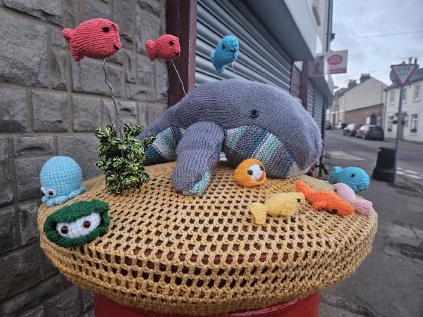 Knitted ocean‑themed post box topper in Higham featuring a large grey whale surrounded by colourful woollen fish, seaweed, and small sea creatures on a crocheted beige cover.