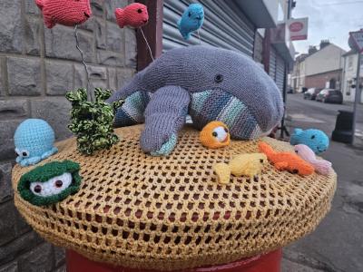 Knitted ocean‑themed post box topper in Higham featuring a large grey whale surrounded by colourful woollen fish, seaweed, and small sea creatures on a crocheted beige cover.