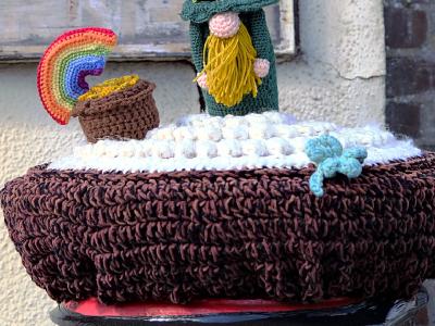 Crocheted post box topper in Shorne Village, Kent showing a leprechaun with yellow beard, pot of gold and rainbow on a textured white and brown base.