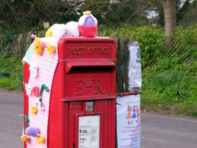 Red village postbox decorated with crocheted Easter designs beside a roadside.