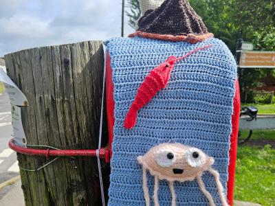 Reverse view of a crochet sea-themed postbox topper. A blue crocheted panel features three red and cream sea creatures: a lobster near the top, a jellyfish with dangling tentacles in the centre, and a crab at the base. A whimsical snail figure with stalk eyes sits atop the installation.
