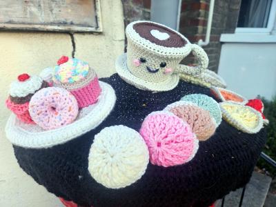 Crochet post box topper in Shorne village depicting a tea-and-cakes theme - a smiling cream-colored teacup with a heart design and pink cheeks sits on a saucer at the top, surrounded by an assortment of crocheted treats on a black knitted base—including a pink cherry-topped cupcake, a rainbow-speckled cupcake, a sprinkle-covered pink donut, pastel-colored macarons, cream and pink meringues, and orange and yellow tart-shaped decorations.