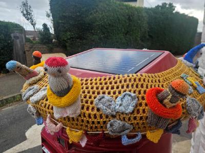 Red UK pillar box covered in mustard-yellow knitted mesh with handmade knitted figurines wearing swimming costumes and bright rubber rings. A solar panel sits on top. Residential street background.